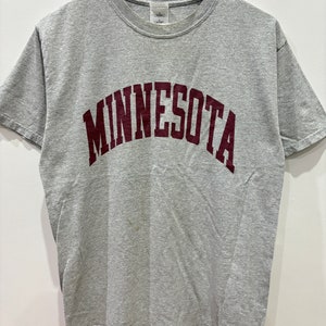 Könnte beinhalten: Ein graues T-Shirt mit einem burgunderfarbenen "Minnesota"-Aufdruck auf der Vorderseite.