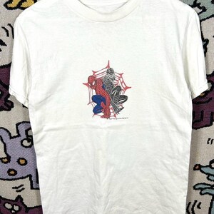 May include: White t-shirt with a Spider-Man graphic in red, blue, and black, set against a red spiderweb. The shirt is displayed on a white hanger. The text "SPIDER-MAN" is visible.
