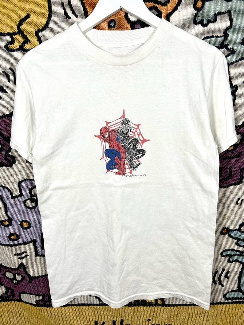 May include: White t-shirt with a Spider-Man graphic in red, blue, and black, set against a red spiderweb. The shirt is displayed on a white hanger. The text "SPIDER-MAN" is visible.