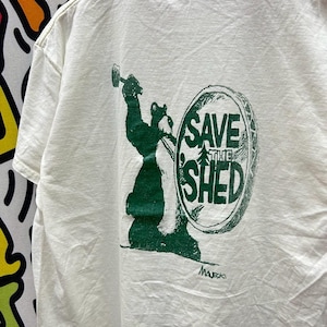 May include: Off-white t-shirt with a green graphic on the back. The graphic features a bear holding a hammer and a shield with the words "SAVE THE SHED" and a tree. The shirt has a crew neck and short sleeves.