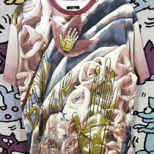 May include: A short-sleeved t-shirt with a unique all-over print featuring abstract shapes, hands, and text. The shirt has a burgundy neckline and sleeve edges. The design incorporates shades of brown, white, and green.