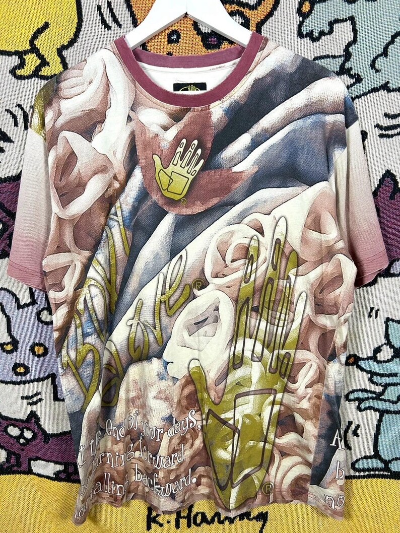 May include: A short-sleeved t-shirt with a unique all-over print featuring abstract shapes, hands, and text. The shirt has a burgundy neckline and sleeve edges. The design incorporates shades of brown, white, and green.