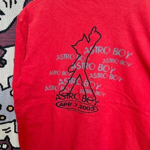 May include: Red t-shirt featuring a graphic of Astro Boy with the text "ASTRO BOY" repeated. The design includes the date "APR. 7, 2003". The shirt is made of a soft material.
