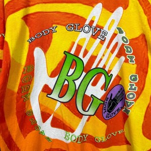 May include: A long-sleeved shirt with a vibrant orange, yellow, and white tie-dye pattern. The shirt features the words "BODY GLOVE" in a circular design around a white handprint and the letters "BG" in green.