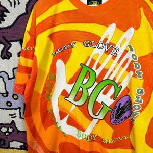 May include: Orange and yellow Body Glove t-shirt with a large white hand graphic. The shirt features the text "BODY GLOVE" in a circular pattern around the hand and the letters "BG" in green. A purple oval with a hand graphic is also present.