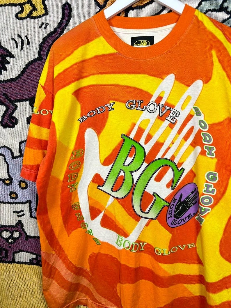May include: Orange and yellow Body Glove t-shirt with a large white hand graphic. The shirt features the text "BODY GLOVE" in a circular pattern around the hand and the letters "BG" in green. A purple oval with a hand graphic is also present.