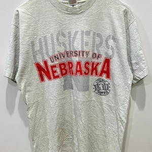 Könnte beinhalten: Ein graues T-Shirt mit einem roten und weißen Grafikdruck, auf dem "Huskers University of Nebraska" steht, darunter ein roter Kreis mit der Aufschrift "Authentic Big Red Sportswear".