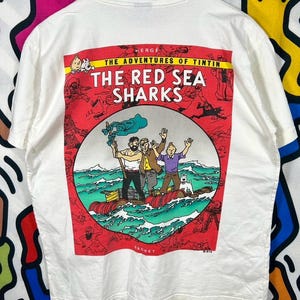 Może przedstawiać: Biały t-shirt z nadrukiem graficznym z Przyg&oacute;d Tintina: Koks w ładowni. Wz&oacute;r przedstawia Tintina, Kapitana Haddoka i Snowy'ego na tratwie na oceanie, otoczonych czerwoną ramką z mniejszymi ilustracjami komiksowymi.