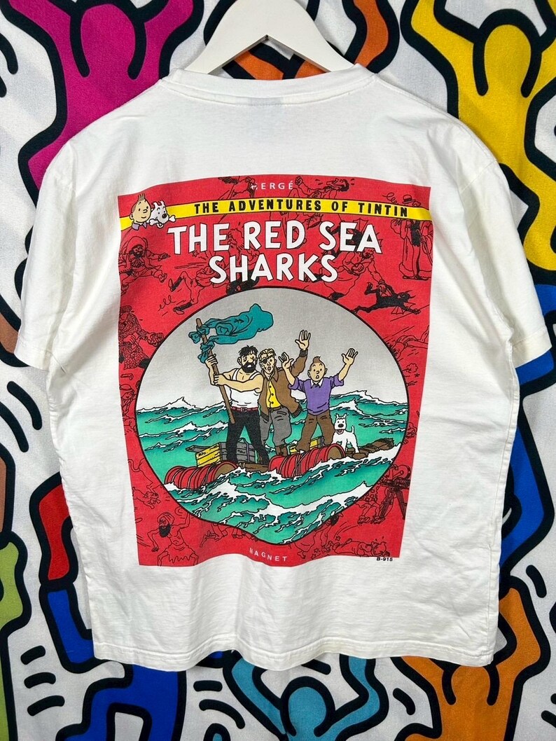 Może przedstawiać: Biały t-shirt z nadrukiem graficznym z Przyg&oacute;d Tintina: Koks w ładowni. Wz&oacute;r przedstawia Tintina, Kapitana Haddoka i Snowy'ego na tratwie na oceanie, otoczonych czerwoną ramką z mniejszymi ilustracjami komiksowymi.