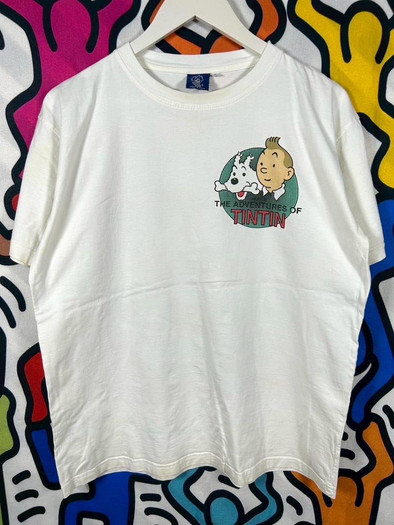 Może przedstawiać: Biały t-shirt z kr&oacute;tkim rękawem z grafiką na piersi przedstawiającą postacie Tintina i Snowy'ego wewnątrz zielonego koła. Poniżej postaci znajduje się napis THE ADVENTURES OF TINTIN czerwonymi i czarnymi literami na białym tle.