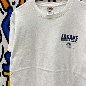 May include: A white t-shirt with the text "ESCAPE FROM L.A." in navy blue, along with the Paramount Pictures logo. The shirt is hanging on a wooden hanger. The shirt is made of a soft material.