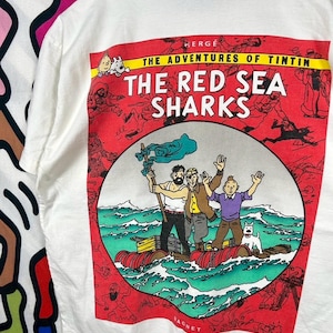 Może przedstawiać: Biały t-shirt z nadrukiem graficznym z serii Przygody Tintina: Koks w ładowni. Wz&oacute;r przedstawia Tintina, Milusia i dw&oacute;ch towarzyszy na tratwie na oceanie, otoczonych czerwoną ramką z r&oacute;żnymi małymi ilustracjami postaci z komiksu.