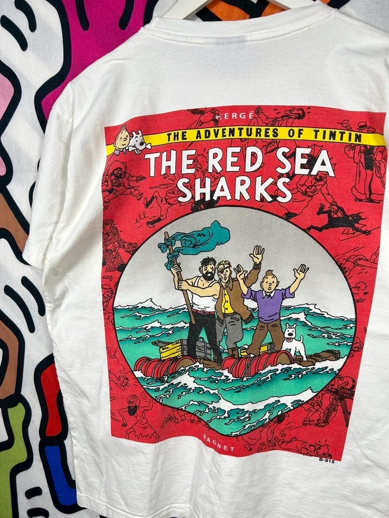 Może przedstawiać: Biały t-shirt z nadrukiem graficznym z serii Przygody Tintina: Koks w ładowni. Wz&oacute;r przedstawia Tintina, Milusia i dw&oacute;ch towarzyszy na tratwie na oceanie, otoczonych czerwoną ramką z r&oacute;żnymi małymi ilustracjami postaci z komiksu.