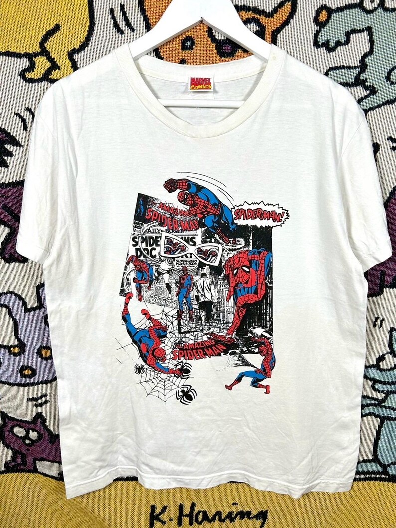 May include: White t-shirt featuring a comic book style graphic of Spider-Man in action poses. The design includes the words "Spider-Man" and "Marvel Comics". The shirt is hanging on a white hanger.