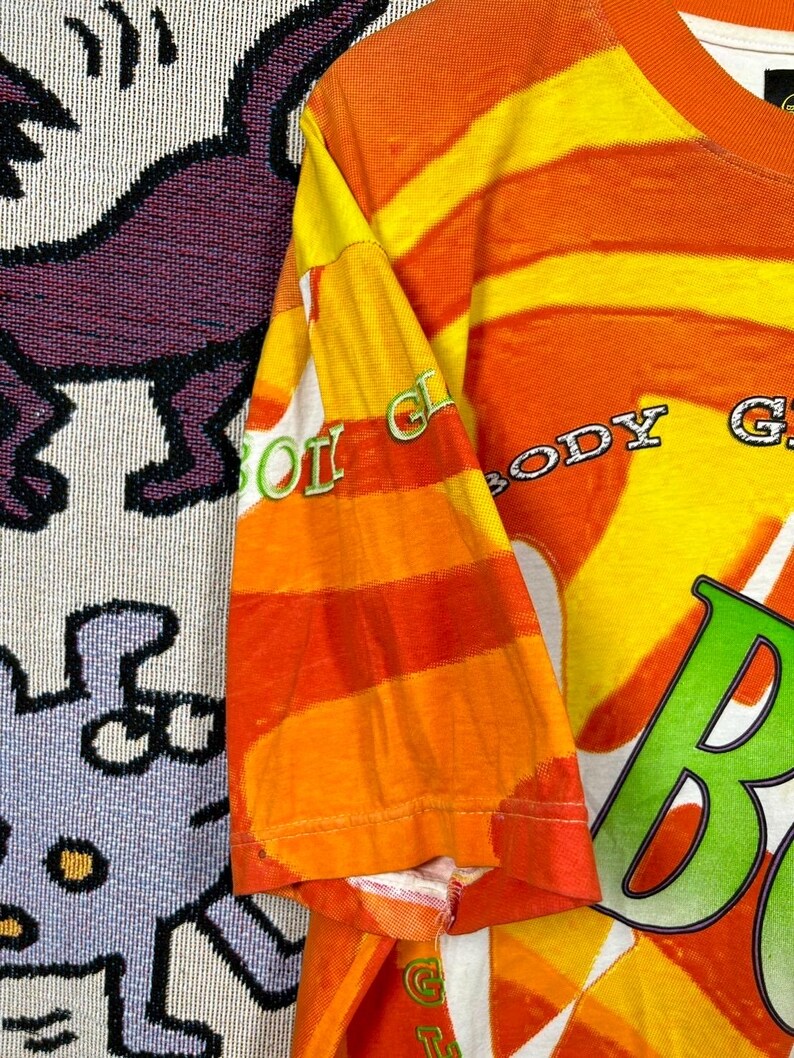 May include: Orange and yellow striped t-shirt with the text "BODY GLOW" in a green and white font. The sleeve has the text "BODY GLOW" in a green and white font. The shirt is made of a soft, comfortable material.