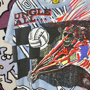 Puede incluir: Camiseta azul claro con un gr&aacute;fico de un jugador de voleibol y las palabras "Jungle Ball". El dise&ntilde;o presenta un bal&oacute;n de voleibol, un patr&oacute;n de cuadros negros y azules y detalles en rojo y amarillo. La camiseta est&aacute; colgada en una percha blanca.