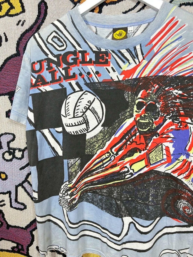 Puede incluir: Camiseta azul claro con un gr&aacute;fico de un jugador de voleibol y las palabras "Jungle Ball". El dise&ntilde;o presenta un bal&oacute;n de voleibol, un patr&oacute;n de cuadros negros y azules y detalles en rojo y amarillo. La camiseta est&aacute; colgada en una percha blanca.