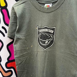 May include: A green t-shirt with a black and white graphic of a shield with the text "Turbonegro" and a black and white illustration of a hat inside the shield.