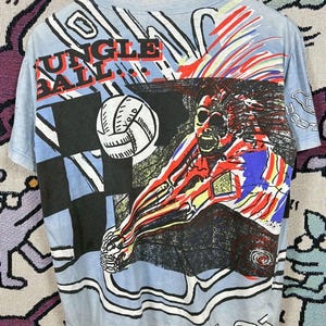 Puede incluir: Camiseta azul claro con un gr&aacute;fico colorido de un jugador de voleibol rematando un bal&oacute;n. La camiseta presenta el texto "JUNGLE BALL" en rojo y negro. El dise&ntilde;o incluye un patr&oacute;n de cuadros y elementos abstractos.