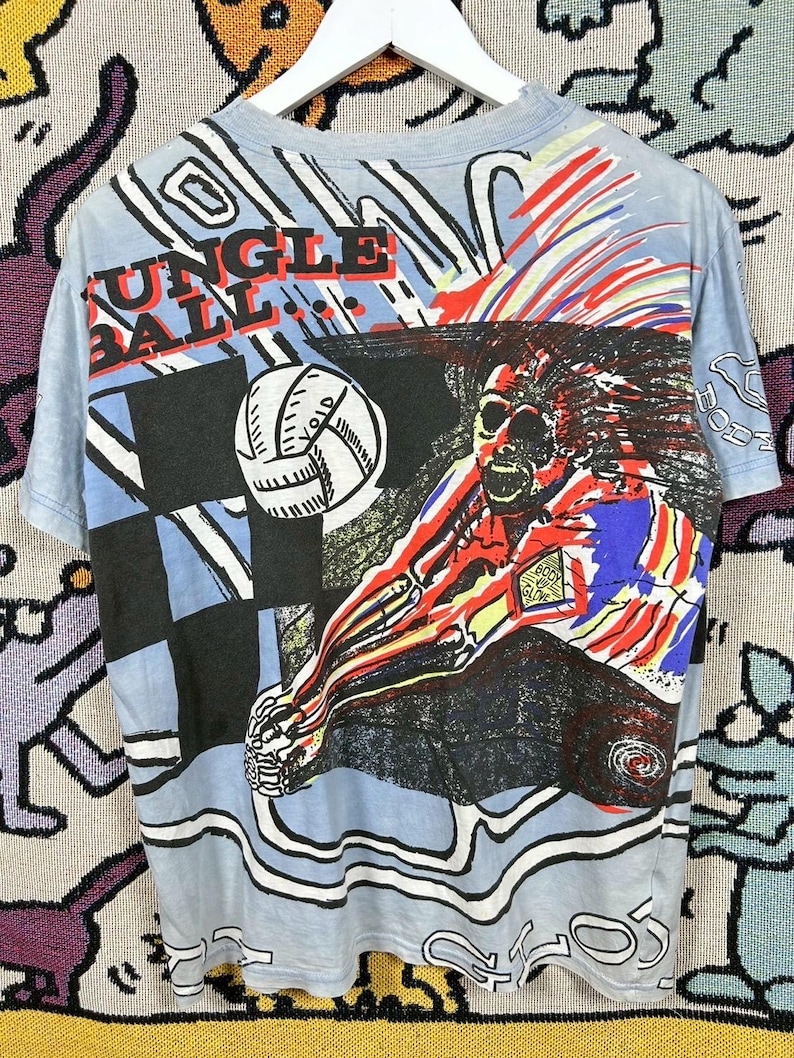 Puede incluir: Camiseta azul claro con un gr&aacute;fico colorido de un jugador de voleibol rematando un bal&oacute;n. La camiseta presenta el texto "JUNGLE BALL" en rojo y negro. El dise&ntilde;o incluye un patr&oacute;n de cuadros y elementos abstractos.