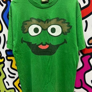 May include: Green t-shirt featuring a large graphic of Oscar the Grouch's face from Sesame Street. The design includes his signature brown eyebrows, white eyes, and a red mouth. The shirt has a crew neck and short sleeves.