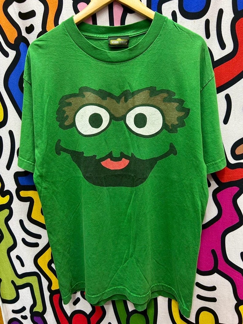 May include: Green t-shirt featuring a large graphic of Oscar the Grouch's face from Sesame Street. The design includes his signature brown eyebrows, white eyes, and a red mouth. The shirt has a crew neck and short sleeves.