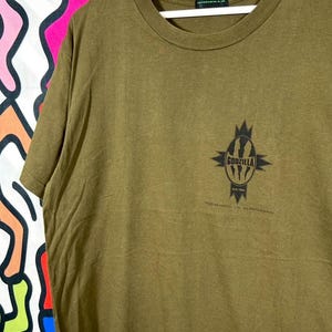 Può includere: Una t-shirt a maniche corte color verde oliva con una grafica nera sul petto. La grafica mostra un emblema ovale stilizzato con il testo Godzilla e New York all'interno, circondato da punte. Una piccola etichetta del marchio Godzilla è sul collo.