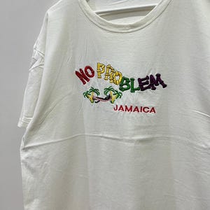 以下が含まれることがあります： 白いTシャツに、カラフルな刺繍で「NO PROBLEM JAMAICA」と書いてあります。デザインにはヤシの木とビーチで横になっている人が描かれています。