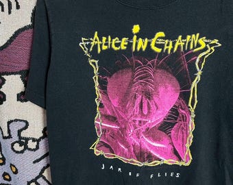 Könnte beinhalten: Schwarzes T-Shirt mit dem Bandnamen "Alice in Chains" in Gelb und einer rosa-schwarzen Fliegen-Grafik. Der Text "Jar of Flies" steht darunter. Das T-Shirt hängt an einem weißen Kleiderbügel.