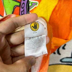 May include: Close-up of a colorful beach towel with the text "BODY GLOVE" and a hand logo. The towel has a vibrant orange, yellow, and purple design. The tag reads "U.S.A." and includes care instructions.
