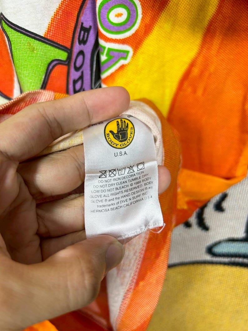 May include: Close-up of a colorful beach towel with the text "BODY GLOVE" and a hand logo. The towel has a vibrant orange, yellow, and purple design. The tag reads "U.S.A." and includes care instructions.