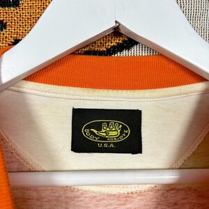 May include: Cream-colored collared shirt with an orange trim and a tag that reads "BODY GLOVE U.S.A." The shirt is hanging on a white hanger. The shirt's collar is orange, and the tag is black with yellow text.