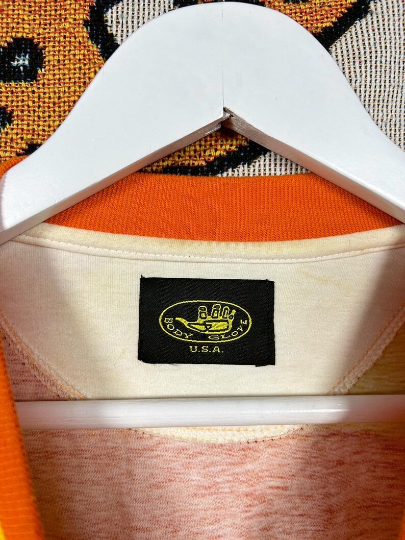 May include: Cream-colored collared shirt with an orange trim and a tag that reads "BODY GLOVE U.S.A." The shirt is hanging on a white hanger. The shirt's collar is orange, and the tag is black with yellow text.