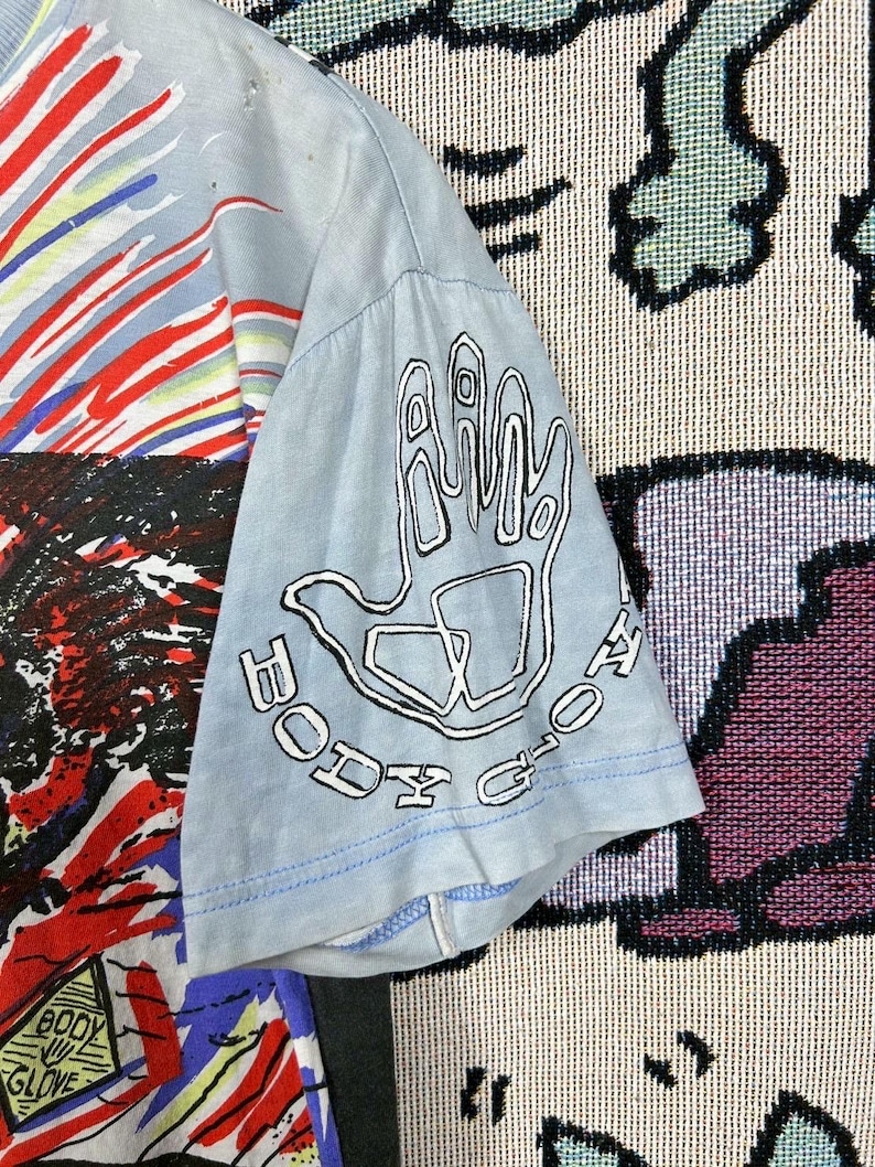 Puede incluir: Camiseta azul claro con un dise&ntilde;o gr&aacute;fico colorido. La manga presenta un contorno blanco de una mano con las palabras "BODY GLOVE" en un dise&ntilde;o circular. La camiseta tiene un aspecto vintage y desgastado.