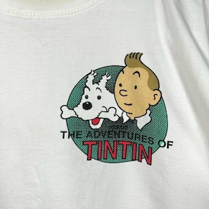 Może przedstawiać: Biały t-shirt z okrągłą grafiką przedstawiającą postać z komiksu Tintina i jego białego psa Milusia. Napis "THE ADVENTURES OF TINTIN" jest wydrukowany czarnymi i czerwonymi literami pod postaciami, na zielonym tle w kropki.