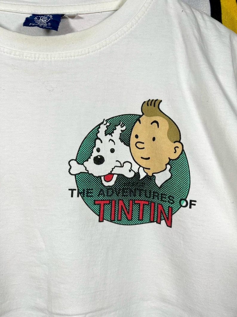 Może przedstawiać: Biały t-shirt z okrągłą grafiką przedstawiającą postać z komiksu Tintina i jego białego psa Milusia. Napis "THE ADVENTURES OF TINTIN" jest wydrukowany czarnymi i czerwonymi literami pod postaciami, na zielonym tle w kropki.