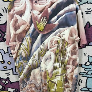 May include: A short-sleeved t-shirt with a complex, abstract design in shades of brown, beige, and green. The shirt features a maroon neckline and a yellow hand logo. Text is visible on the shirt, including the words "The End" and "falling forward."