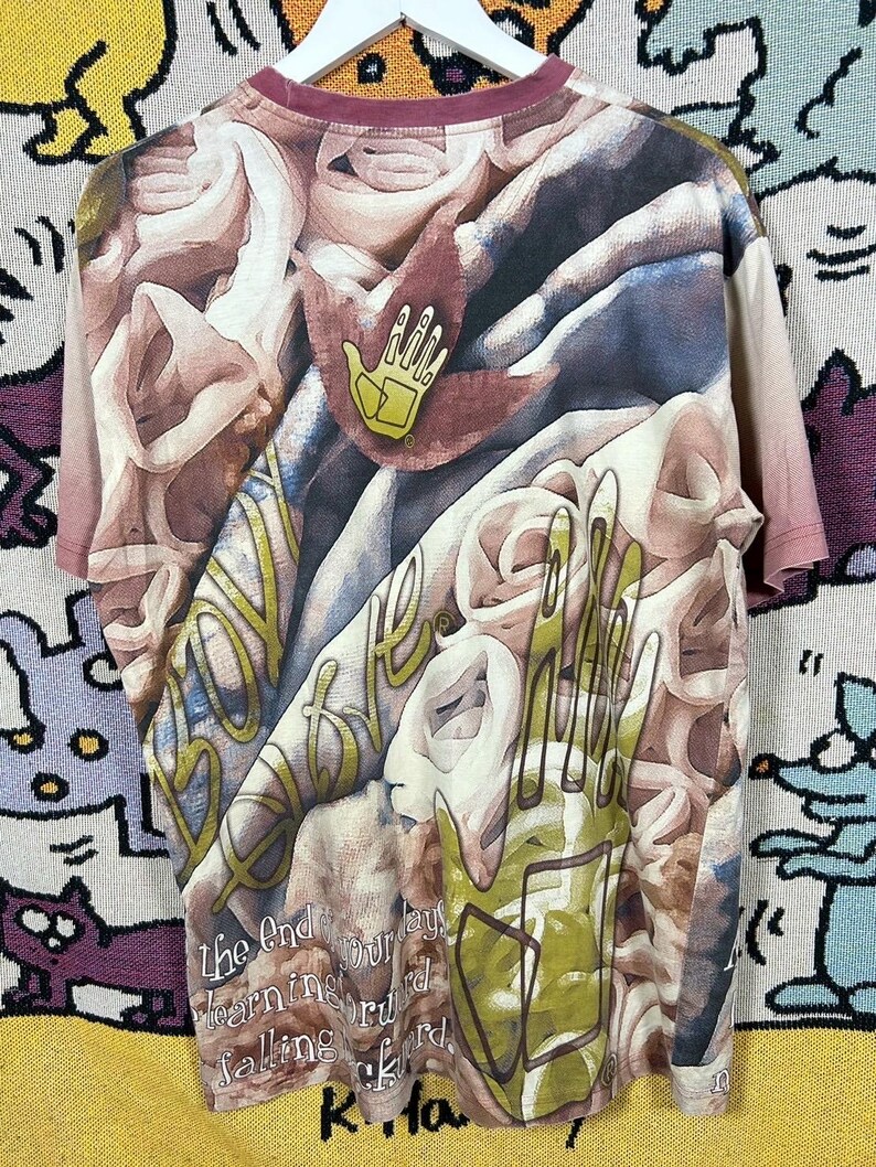May include: A short-sleeved t-shirt with a complex, abstract design in shades of brown, beige, and green. The shirt features a maroon neckline and a yellow hand logo. Text is visible on the shirt, including the words "The End" and "falling forward."