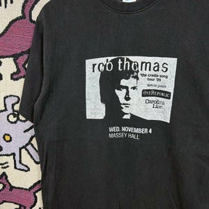 Könnte beinhalten: Schwarzes T-Shirt mit einer Grafik von Rob Thomas und Tour-Informationen. Das Design enthält "the cradle song tour '09", "OneRepublic" und "Carolina Liar". Das Datum "WED. NOVEMBER 4" und "MASSEY HALL" sind ebenfalls aufgedruckt.