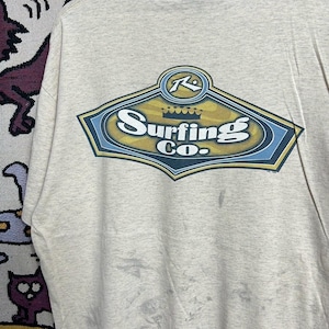 May include: A light beige t-shirt featuring a large graphic on the back that reads "Surfing Co." in a stylized font. The graphic is framed by a blue and gold design. The shirt has a crew neck and short sleeves.