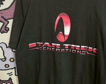 May include: Black Star Trek Generations t-shirt. The front features a red and white Star Trek emblem above the words "STAR TREK GENERATIONS" in red. The shirt is hanging on a white hanger.