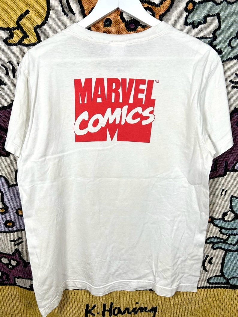 May include: White t-shirt featuring the Marvel Comics logo in red and white. The logo is centered on the back of the shirt. The shirt is hanging on a white hanger.