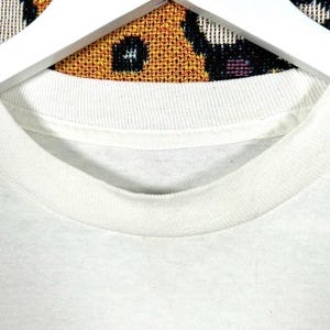 May include: Close-up of a white crew neck t-shirt. The collar is ribbed and the fabric appears to be a soft cotton blend. The shirt is hanging on a white wooden hanger against a patterned background.