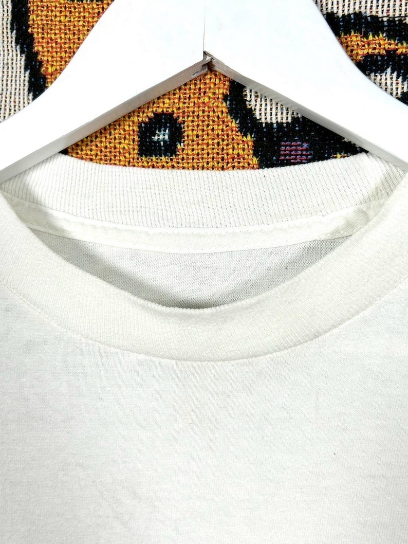 May include: Close-up of a white crew neck t-shirt. The collar is ribbed and the fabric appears to be a soft cotton blend. The shirt is hanging on a white wooden hanger against a patterned background.