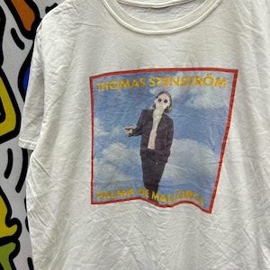 Könnte beinhalten: Weißes T-Shirt mit einer Grafik einer Person mit Sonnenbrille, dem Text "THOMAS STENSTRÖM" und "PALMA DE MALLORCA" in einem roten und gelben Rahmen. Das Bild ist vor einem blauen Himmelshintergrund platziert.