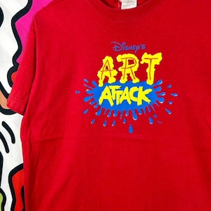 May include: A bright red short-sleeved t-shirt featuring the text Disney's ART ATTACK in yellow letters centred on the chest. The graphic includes a blue paint splatter design behind the yellow text, with a small trademark symbol at the bottom.