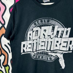 Puede incluir: Una camiseta negra de manga corta con un estampado gráfico en blanco. El diseño incluye un emblema circular con el texto OCALA, A DAY TO REMEMBER y FLORIDA, acompañado por una silueta del estado de Florida en la esquina inferior derecha.