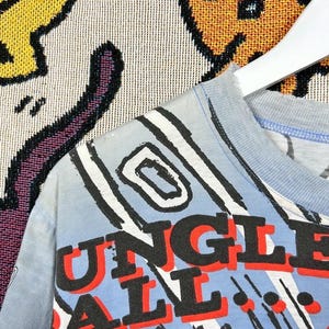 Puede incluir: Camiseta azul claro con las palabras "JUNGLE BALL" en letras rojas y negras. La camiseta presenta un dise&ntilde;o gr&aacute;fico con l&iacute;neas y formas en blanco y negro sobre un fondo azul claro.