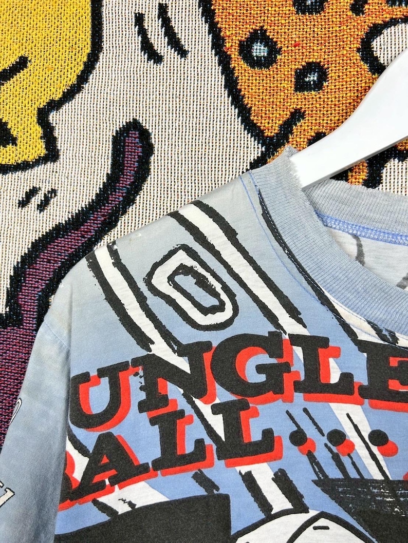 Puede incluir: Camiseta azul claro con las palabras "JUNGLE BALL" en letras rojas y negras. La camiseta presenta un dise&ntilde;o gr&aacute;fico con l&iacute;neas y formas en blanco y negro sobre un fondo azul claro.
