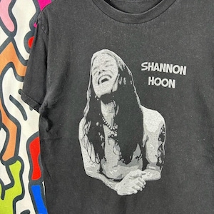 Puede incluir: Una camiseta negra de manga corta con un estampado gráfico gris de Shannon Hoon con el pelo largo y un collar. El texto SHANNON HOON está impreso en letras mayúsculas grises a la derecha del retrato. La camiseta tiene cuello redondo.